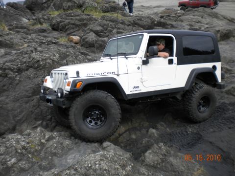 2005 Jeep Wrangler - A white Jeep Wrangler Rubicon navigates a rocky terrain, with a red vehicle visible in the background.