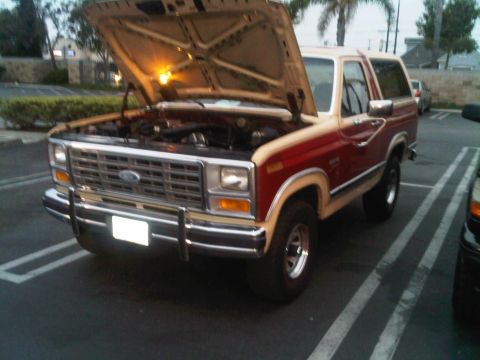 1986 Ford Bronco - The front end of a classic Ford Bronco is visible with its hood raised, revealing the engine compartment.