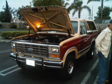 1986 Ford Bronco - This two-tone Ford Bronco has its hood open, revealing the engine bay. A person stands nearby in a parking lot.