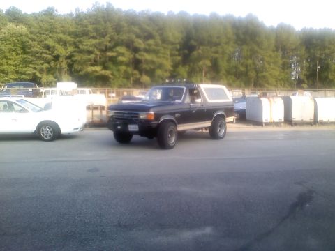 1990 Ford Bronco - A black Ford Bronco with a white removable top is parked in an outdoor lot.
