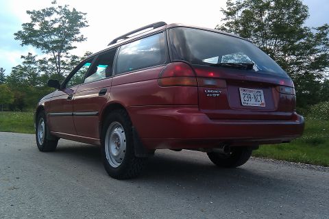 1998 Subaru Legacy - Rear view of a red 1998 Subaru Legacy wagon parked on a paved road with trees in the background.