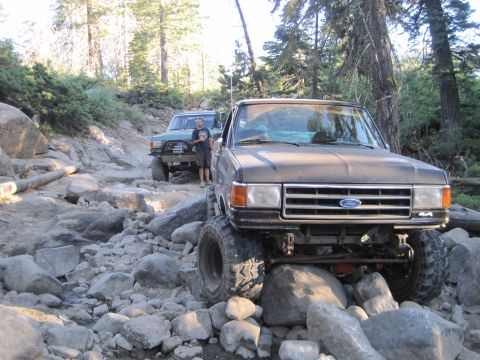 1989 Ford Bronco - Two modified 4x4 trucks tackle a challenging rocky path in a wooded area.