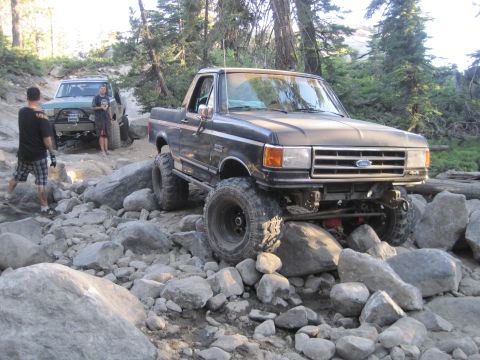1989 Ford Bronco - Two off-road vehicles tackle a challenging rocky path in a wooded area.