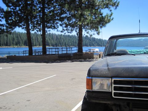 1989 Ford Bronco - A dark gray Ford F-Series truck is parked in a lot overlooking a blue lake surrounded by a dense forest.