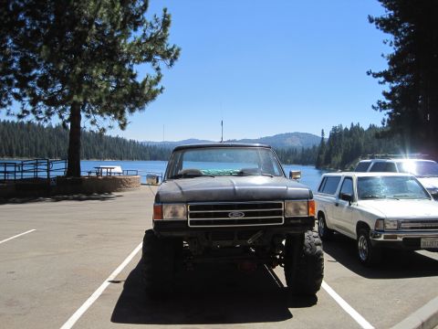1989 Ford Bronco - A lifted black Ford Bronco is parked in a lot with a lake and trees in the background.