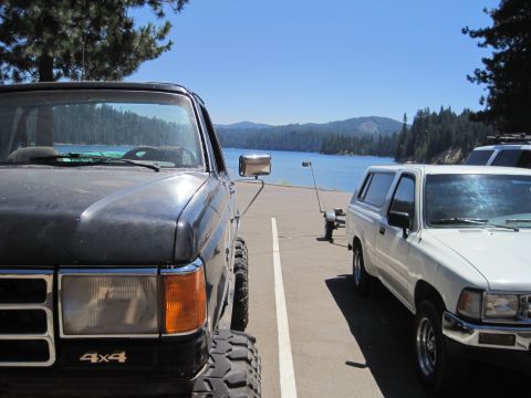 1989 Ford Bronco - A dark 1987 Ford F-Series pickup truck is parked next to a white SUV with a lake in the background.
