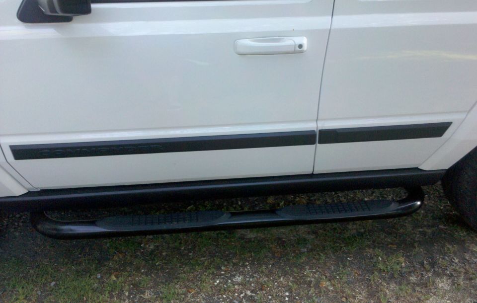 White Jeep Commander side doors with black molding and black tubular step bar over gravel