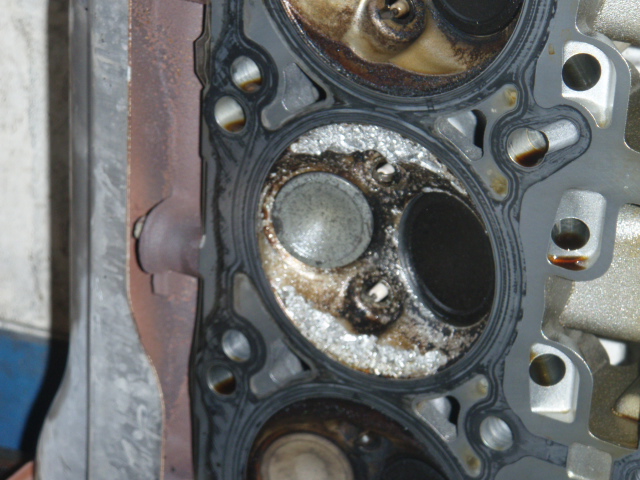 Engine cylinder head with damaged valves and gasket, showing carbon buildup and corrosion