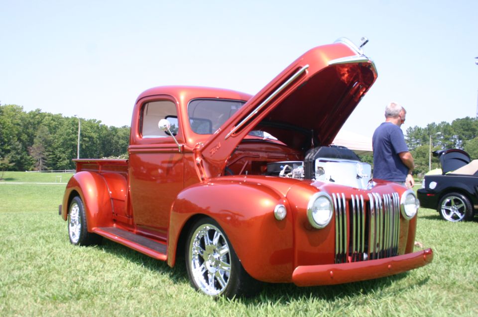 Orange hot rod pickup truck with hood open, showing engine. Guy standing nearby. Black car in background