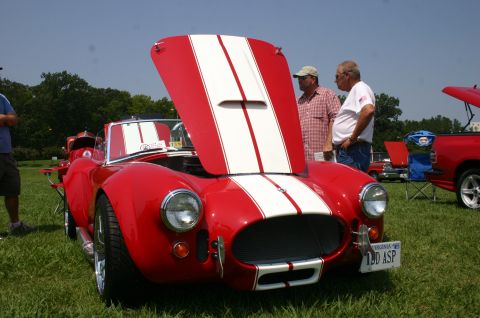 1968 Shelby GT500 - A red AC Cobra with racing stripes is displayed with its hood open at an outdoor car show.