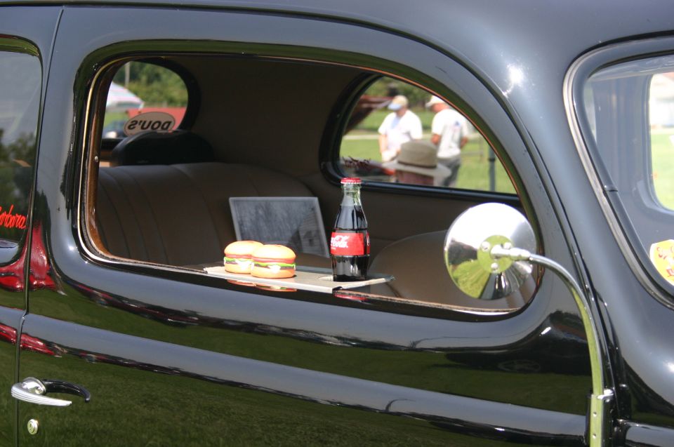 Vintage car interior showing two cheeseburgers and a Coca-Cola bottle on a tray, with people visible through the window