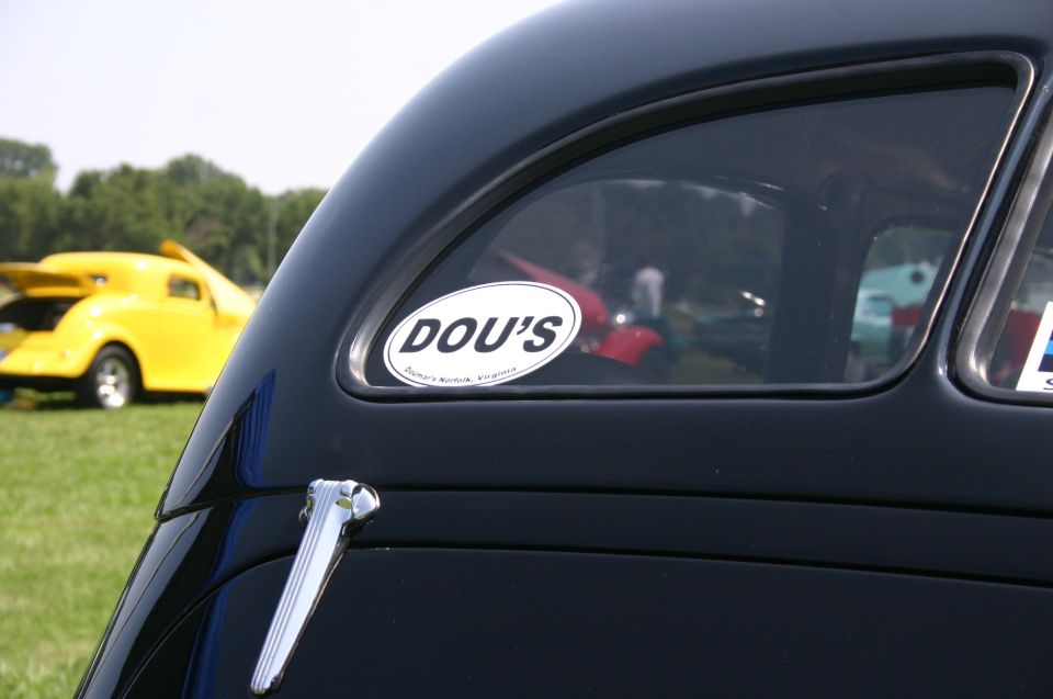 Black car's rear window with a 'DOU'S' sticker. Yellow hot rod in background