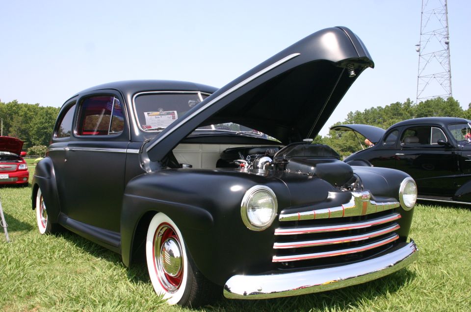 Black coupe with hood open, showing engine. Whitewall tires, chrome grille. Another black coupe visible behind