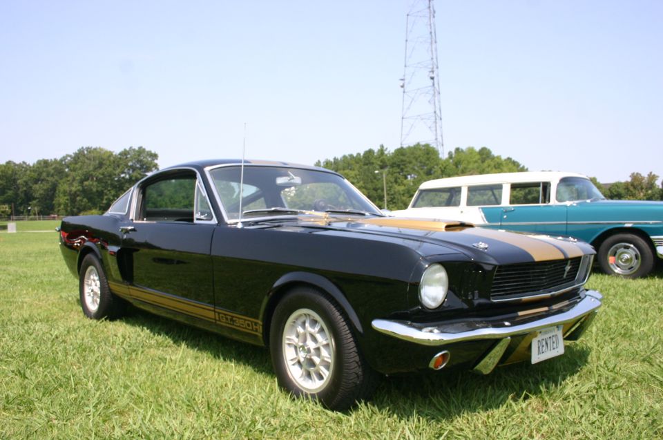 Black 1967 Shelby GT500 with gold stripes parked on grass, another car in background