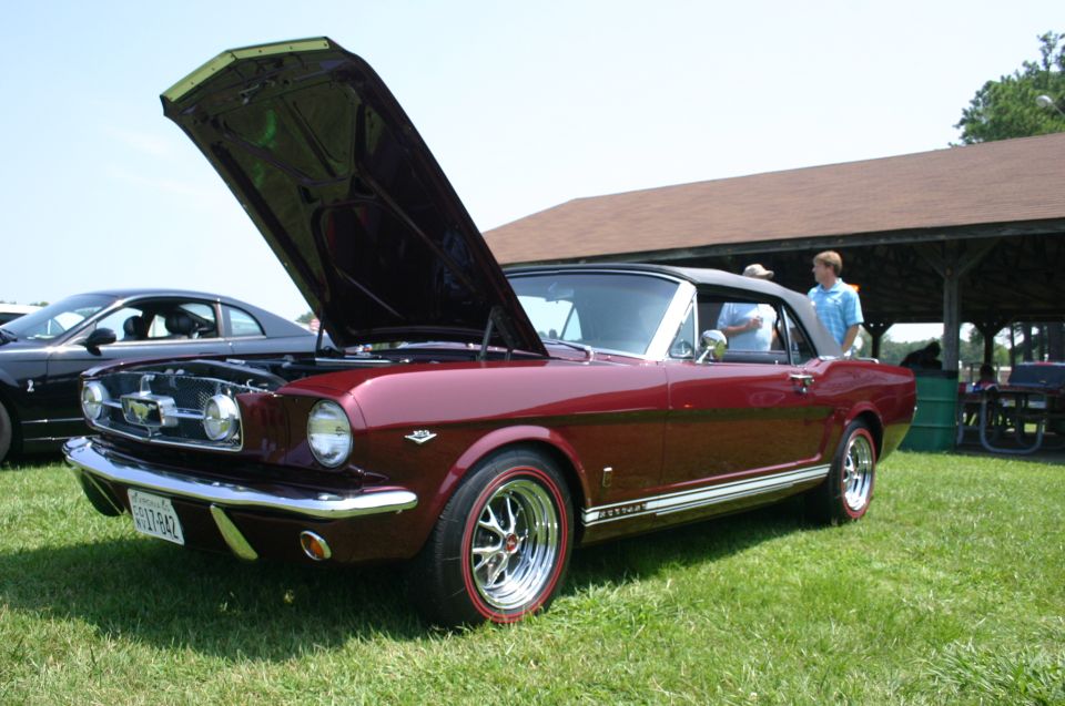 Red 1965 Ford Mustang convertible with hood open, parked on grass next to a black car