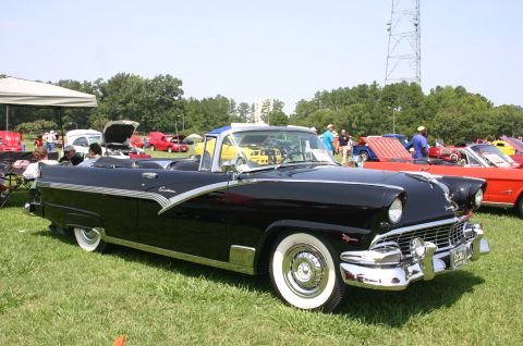 1968 Shelby GT500 - A black 1956 Ford Fairlane convertible is displayed at a car show with other vehicles in the background.