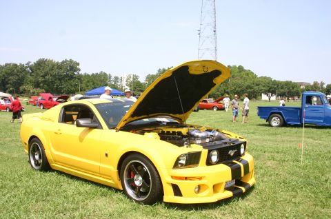 1968 Shelby GT500 - A yellow Ford Mustang with black racing stripes is displayed with its hood open at an outdoor car show.