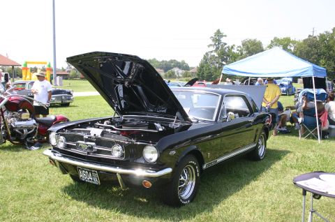1968 Shelby GT500 - A black 1966 Ford Mustang coupe is displayed with its hood open at a car show, with other vehicles and people in the background.