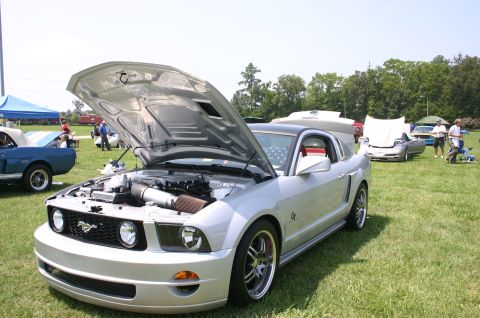 1968 Shelby GT500 - A silver Ford Mustang is displayed with its hood open at a car show, with other vehicles and people in the background.