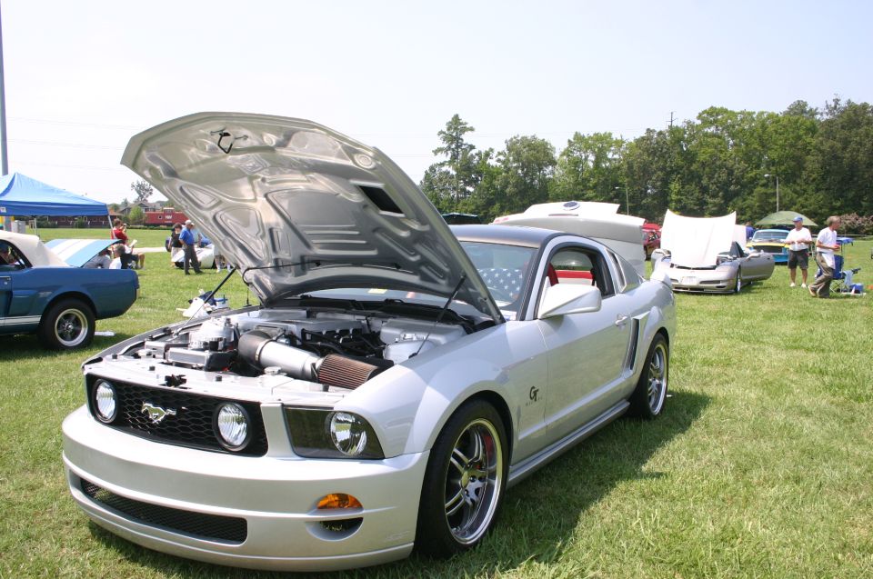 Silver Ford Mustang with hood open, parked on grass at an outdoor car show. Another blue Mustang is visible to the left