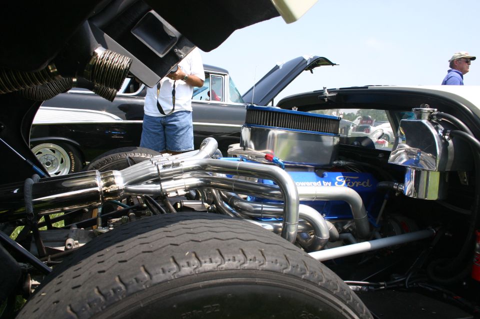 Close-up of a blue Ford V8 engine with chrome headers and air cleaner. Another car is visible in the background