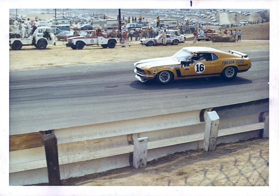 Yellow race car with number 16 and 'FOULGER FORD' text on track, other cars and tow trucks in background