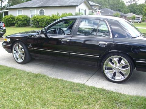 1995 Mercury Grand Marquis - A black sedan with custom chrome wheels is parked in front of a house.