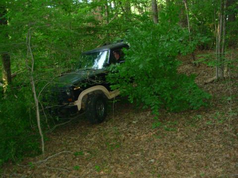 1983 Ford Bronco - A Jeep Cherokee XJ navigates a wooded trail, its front end visible through dense green bushes.