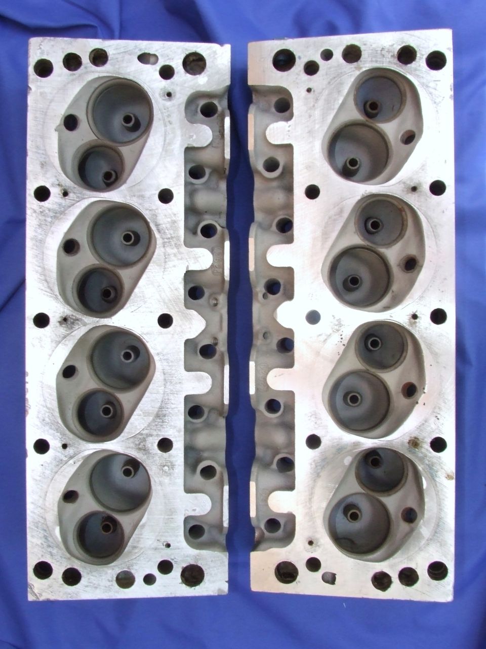 Two aluminum engine cylinder heads with intake and exhaust ports, laid flat on a blue surface