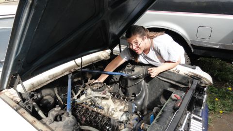 1964 Ford Fairlane - A person works on the engine of a car, with another vehicle parked behind it.