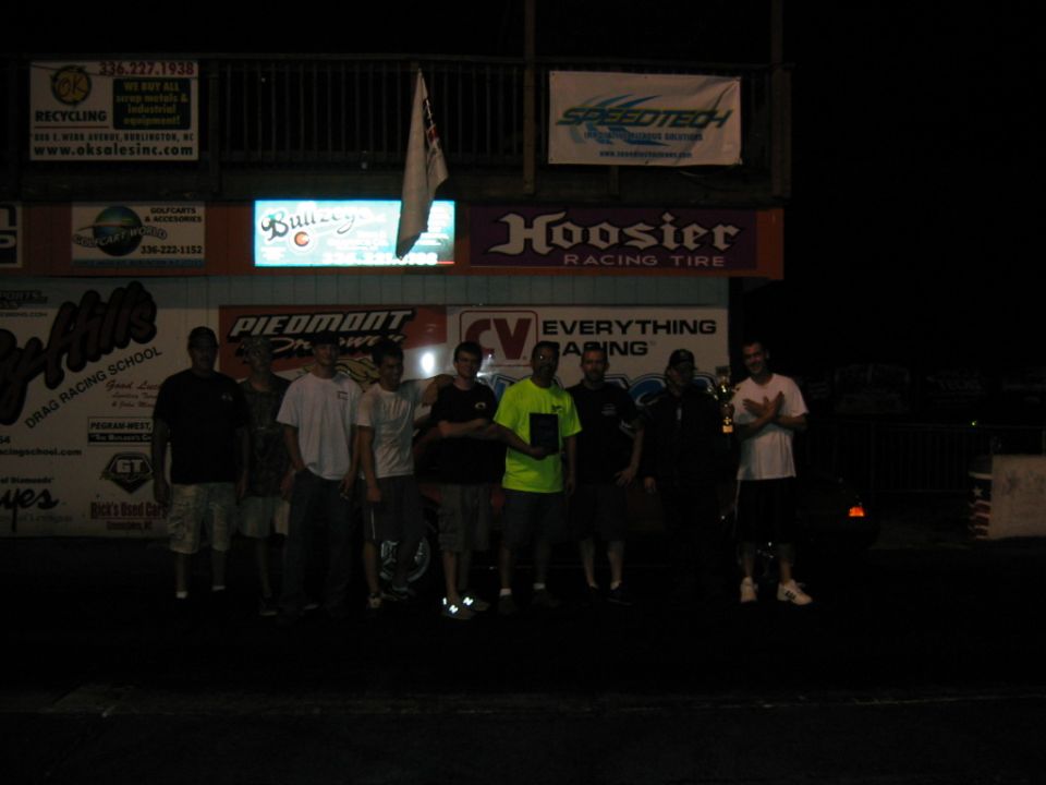 group of guys standing in front of drag strip signs at night, one holding a trophy