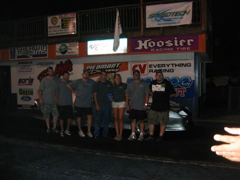 1992 Ford Mustang - A group of people pose in front of a silver car at a race track, with various sponsor banners in the background.