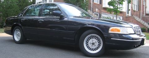 1999 Ford Crown Victoria - A black sedan with a distinctive chrome grille and wire wheels is parked on a residential street.