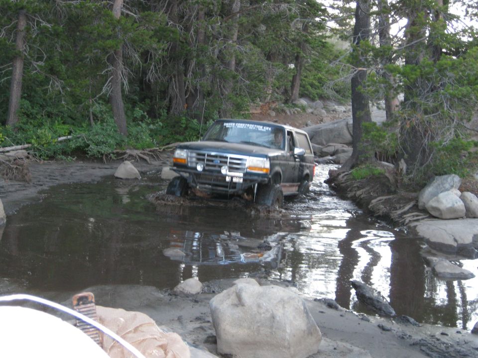 Black Ford Bronco driving through muddy water, front bumper modified with lights