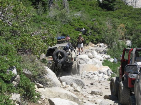 1986 Ford Bronco - Two off-road vehicles traverse a challenging, boulder-strewn path in a wooded area.
