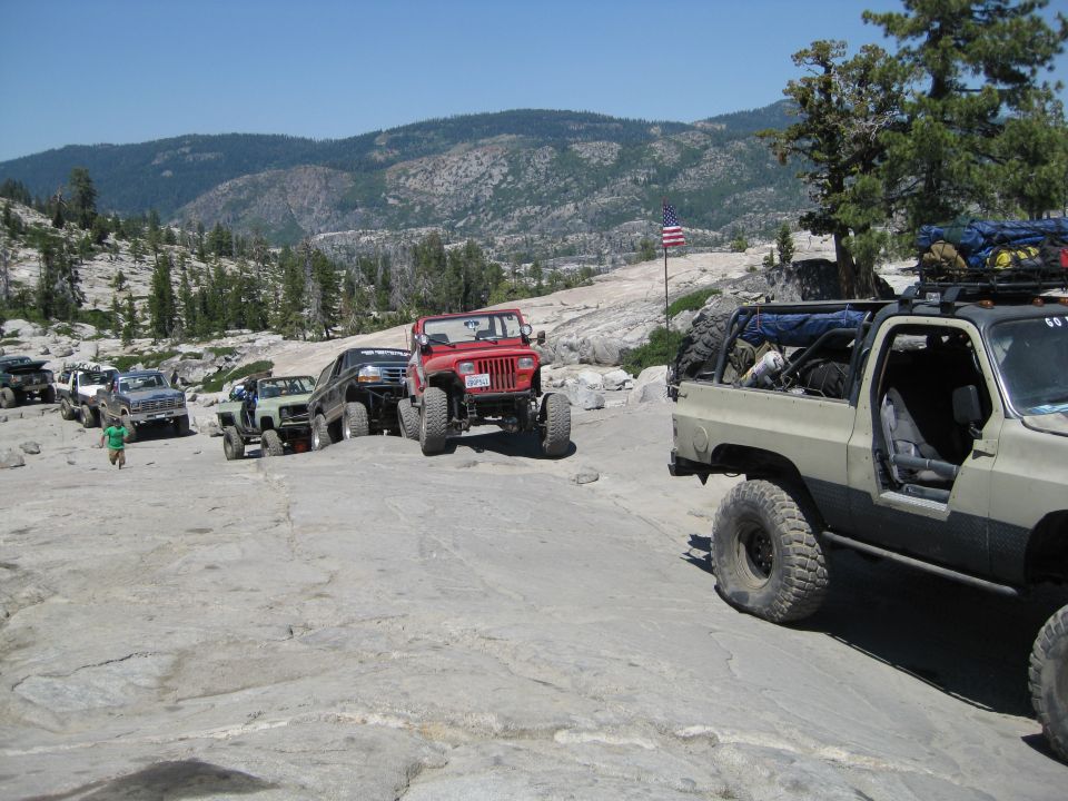 Several off-road vehicles, including a red Jeep and a tan SUV, navigate a rocky, mountainous landscape