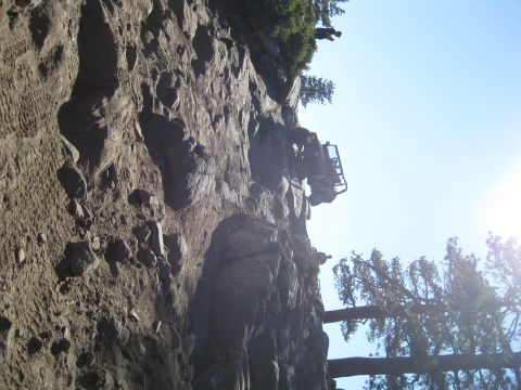 1986 Ford Bronco - An off-road vehicle is precariously positioned on a sheer rock face, with trees and a bright sun visible in the background.