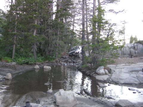 1986 Ford Bronco - An off-road vehicle carefully maneuvers through a challenging rocky terrain with water and trees.