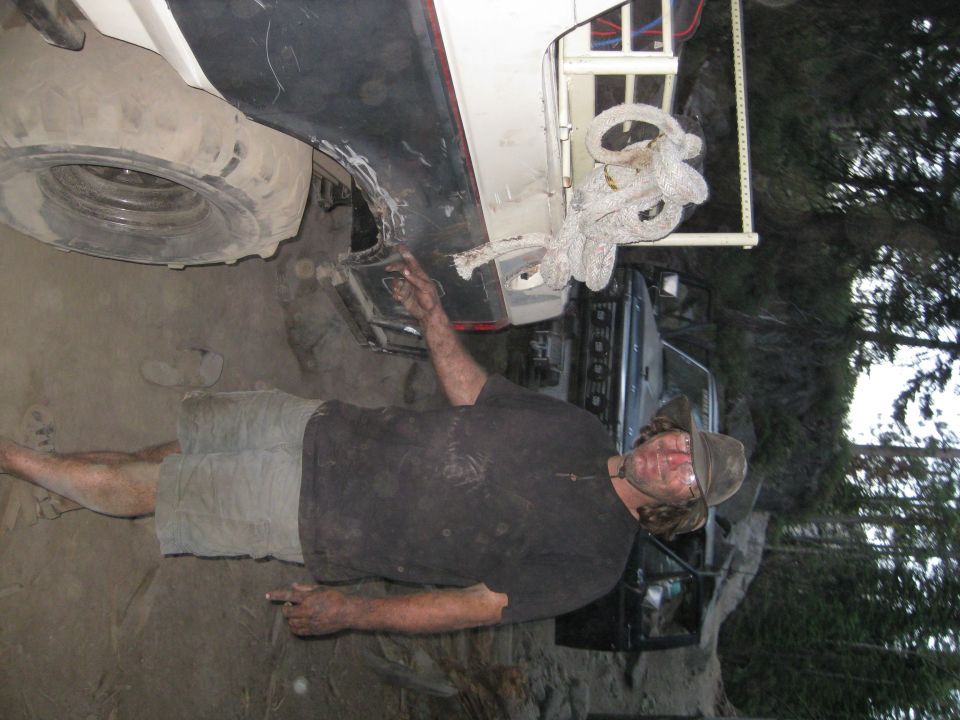 Man with dirty hands and a hat works on the underside of a white vehicle, with a spare tire visible. Another vehicle is partially seen behind him