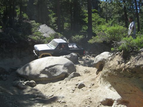 1986 Ford Bronco - A dark blue pickup truck navigates a challenging, rocky off-road trail surrounded by trees and boulders.