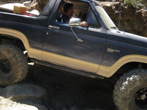 1986 Ford Bronco - A Ford Bronco is shown off-roading over rocks, with a driver visible inside.
