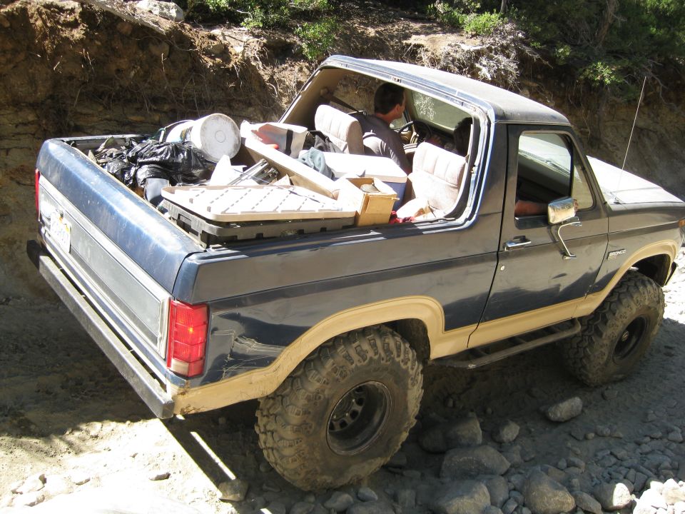 Dark blue Ford Bronco with tan trim, loaded with items in the bed and interior, driving on a rocky trail