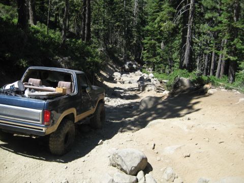 1986 Ford Bronco - This Ford Bronco navigates a challenging, rocky path through a dense forest, with another vehicle ahead.