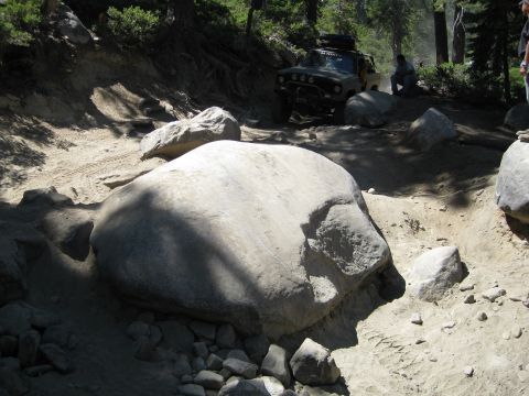 1986 Ford Bronco - An off-road vehicle navigates a challenging rocky trail with trees and boulders surrounding it.