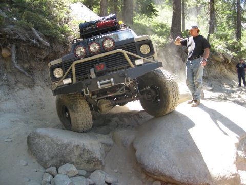 1986 Ford Bronco - A modified off-road vehicle tackles a challenging rocky trail with spectators watching.