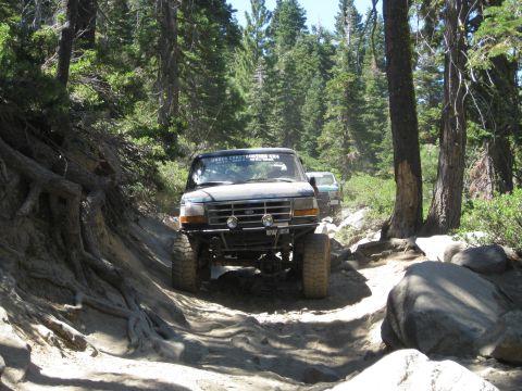 1986 Ford Bronco - Two off-road vehicles navigate a challenging, wooded trail with large rocks and exposed tree roots.