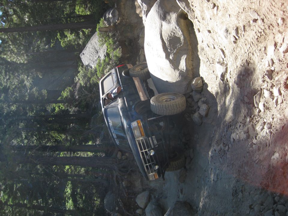1986 Ford Bronco tilted on its side on a narrow, rocky trail with trees in the background