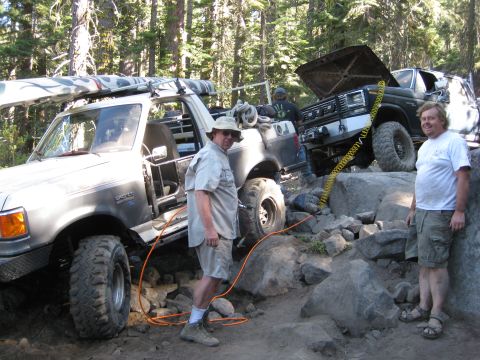 1986 Ford Bronco - Two off-road trucks navigate a rocky trail in a forest setting.