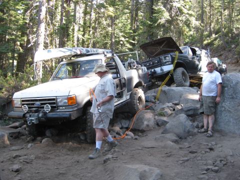 1986 Ford Bronco - Two off-road vehicles are shown on a challenging rocky path in a wooded area, with one vehicle carrying a canoe.