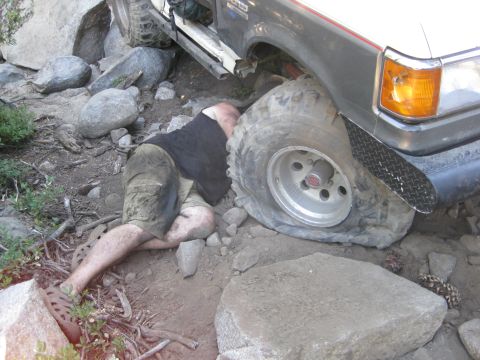 1986 Ford Bronco - A person is stuck with a flat tire on a rocky trail, with the vehicle partially on top of them.